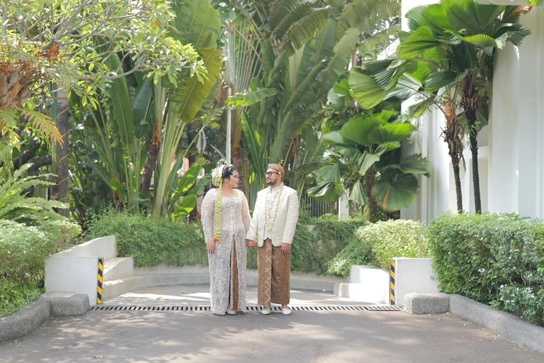 Foto 03: Pasangan pengantin berbusana adat berdiri bergandengan tangan di taman dengan pepohonan tropis — 20230715-P1139979.jpg